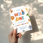 Hand holding a colorful 'Wild One' birthday invitation card with animal illustrations.